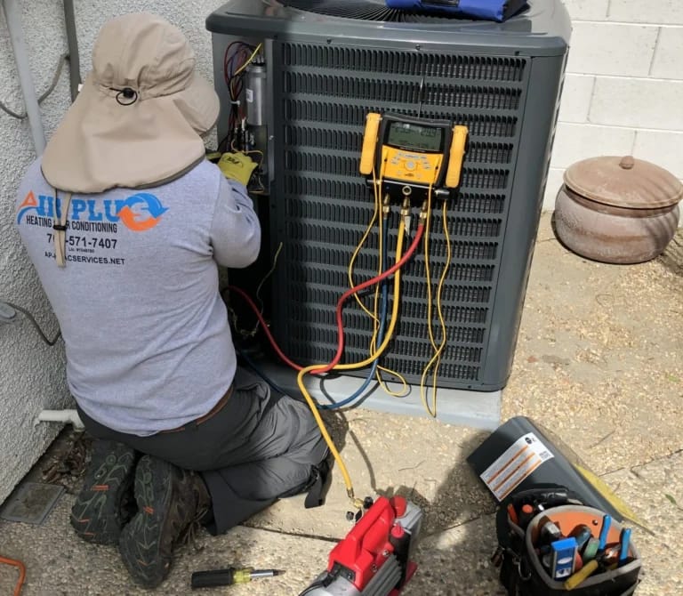 Air Plus technician checking refrigerant operating pressures on an outdoor AC condenser with digital manifold gauges.