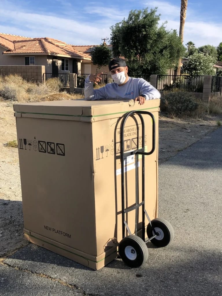 Air Plus HVAC technician posing behind a large packaged unit on a hand truck outside a residential neighborhood.