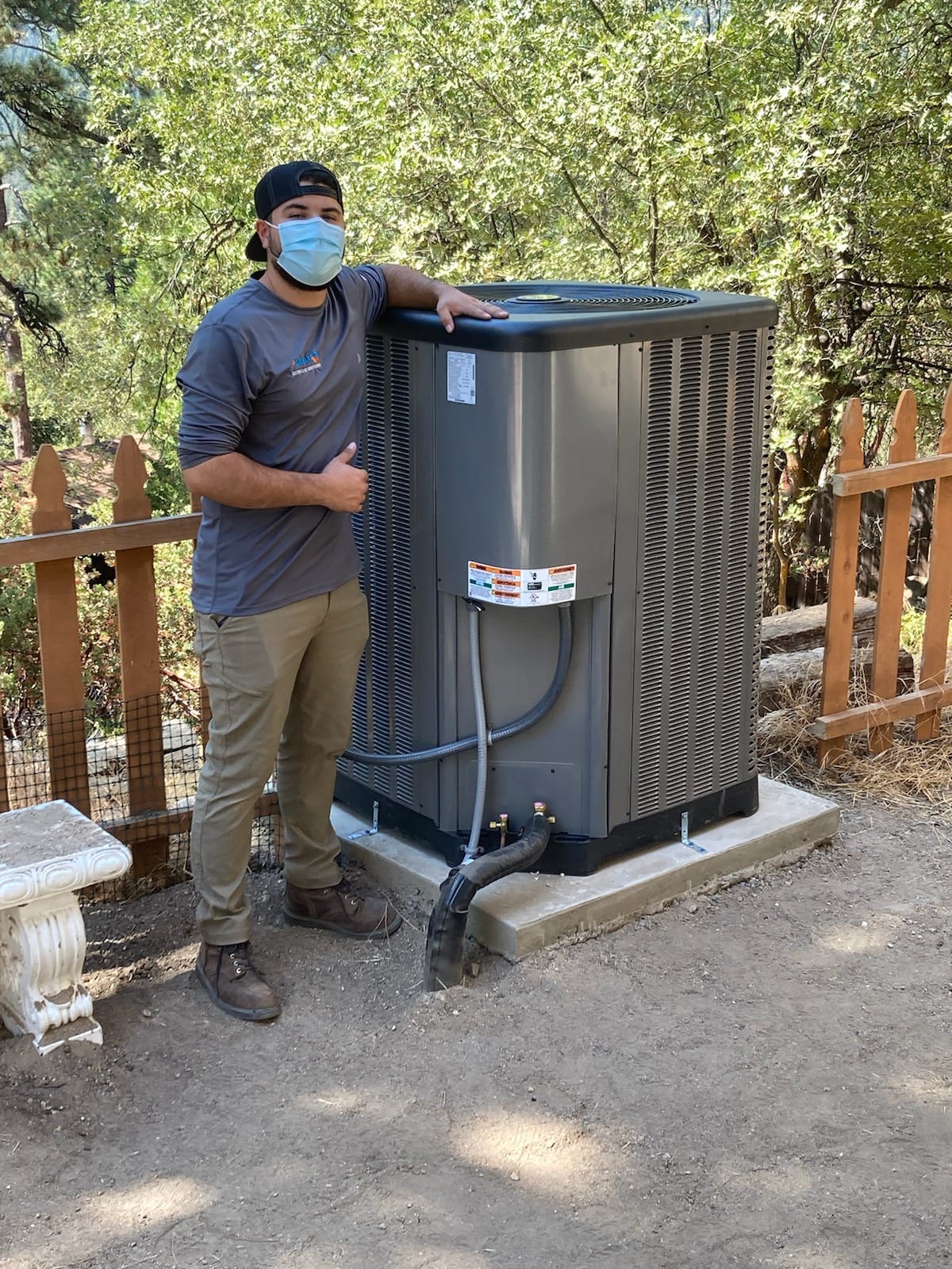 Air Plus HVAC technician giving two thumbs up while servicing an outdoor central air conditioning unit in a residential yard.