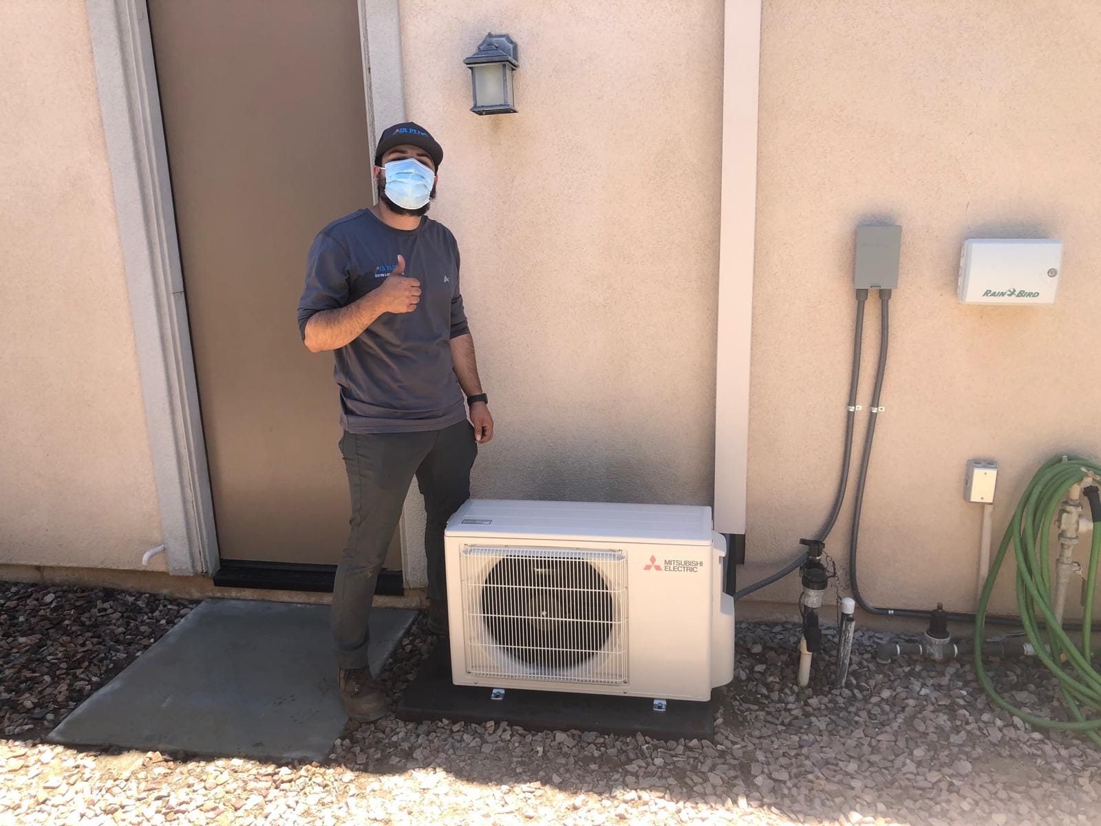 HVAC technician giving a thumbs up beside a newly installed Mitsubishi Electric outdoor mini-split heat pump unit outside a home.
