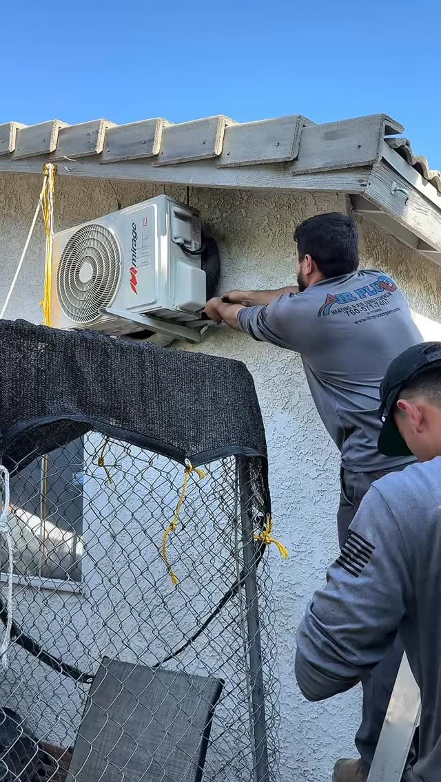 Professional HVAC technicians installing a wall-mounted Mirage mini-split heat pump air conditioning unit on a home exterior.