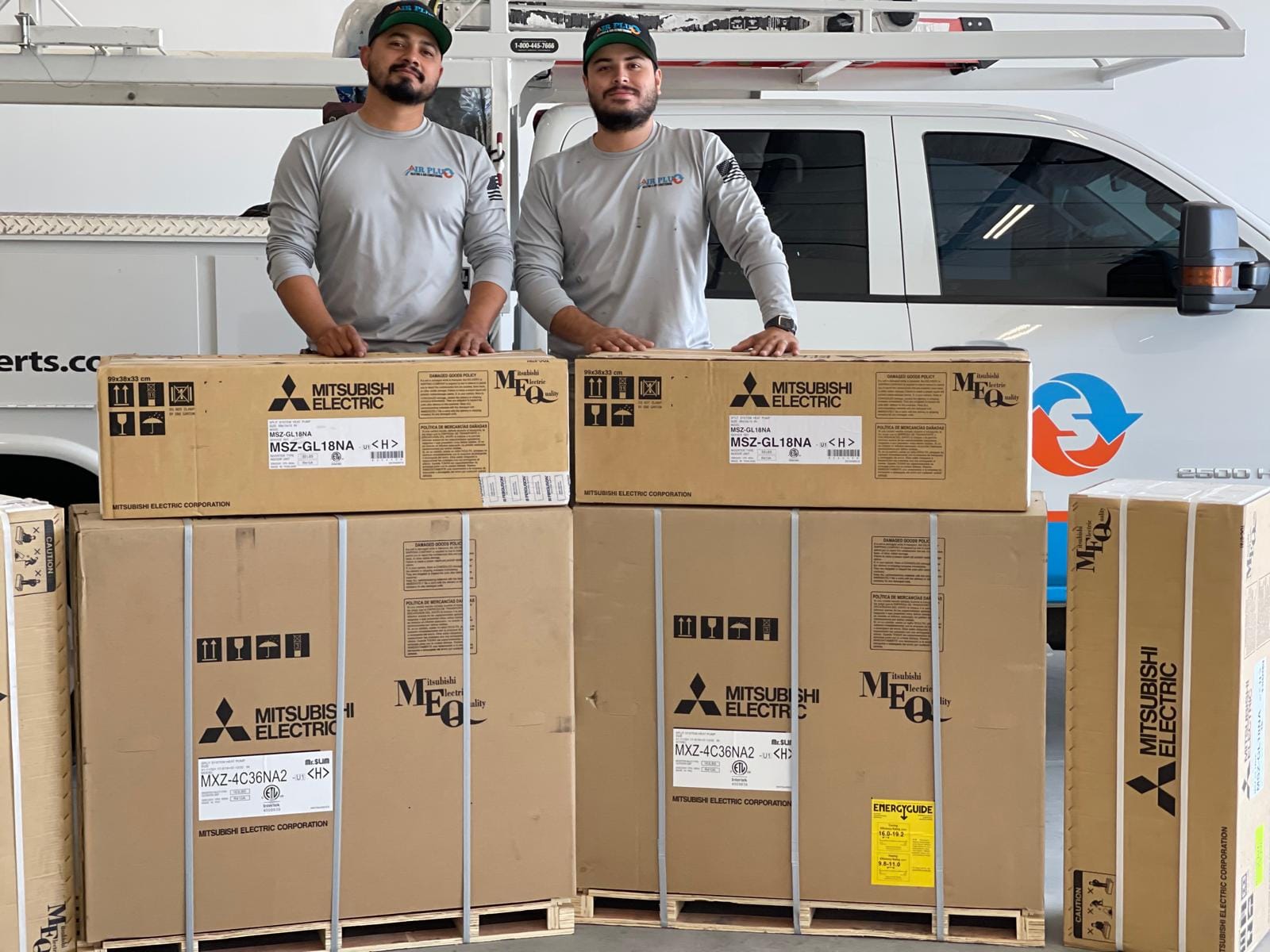 Two Air Plus HVAC technicians posing with packaged Mitsubishi Electric split AC units in front of a service truck, ready for installation.